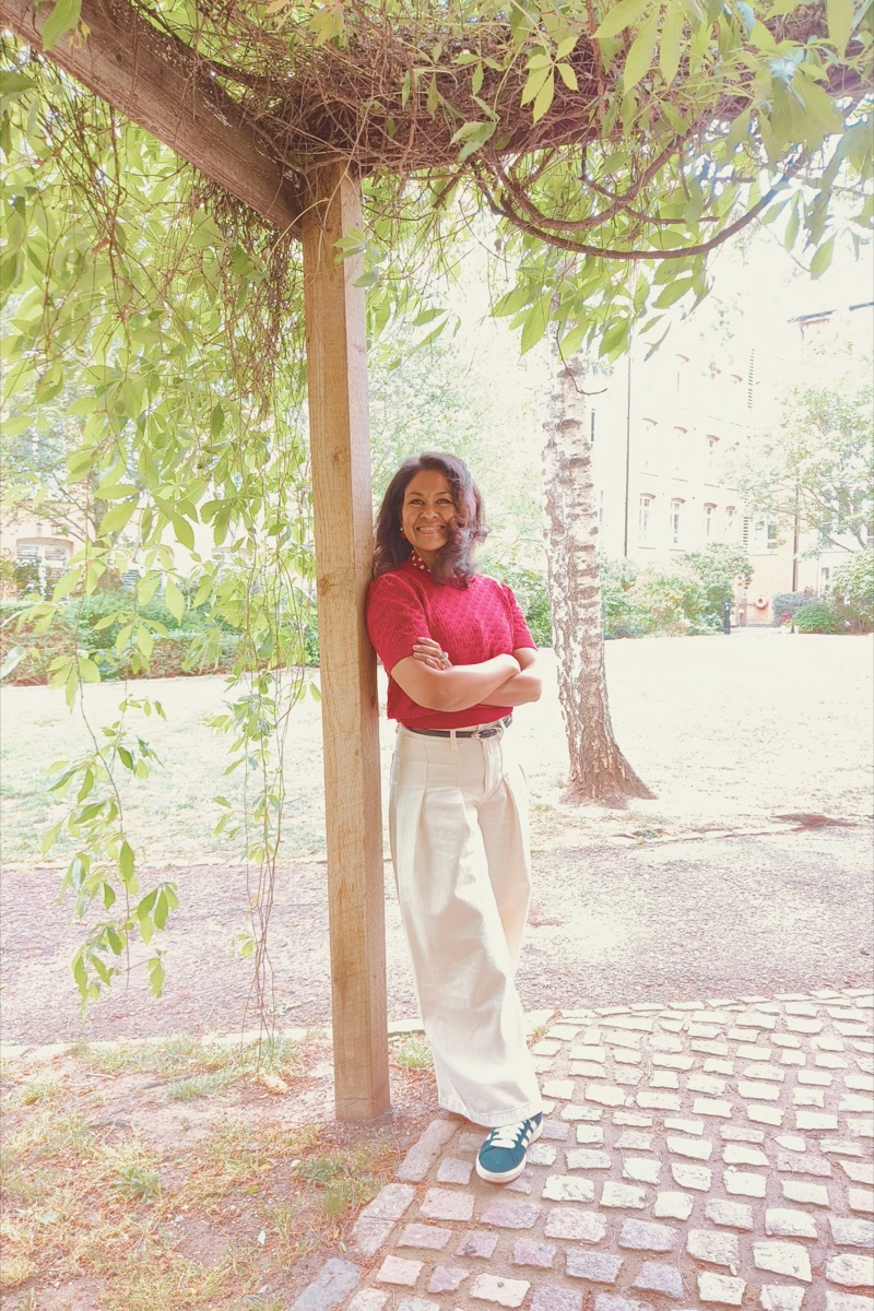 Natasha leaning against a wooden post in a leafy garden, arms folded, smiling